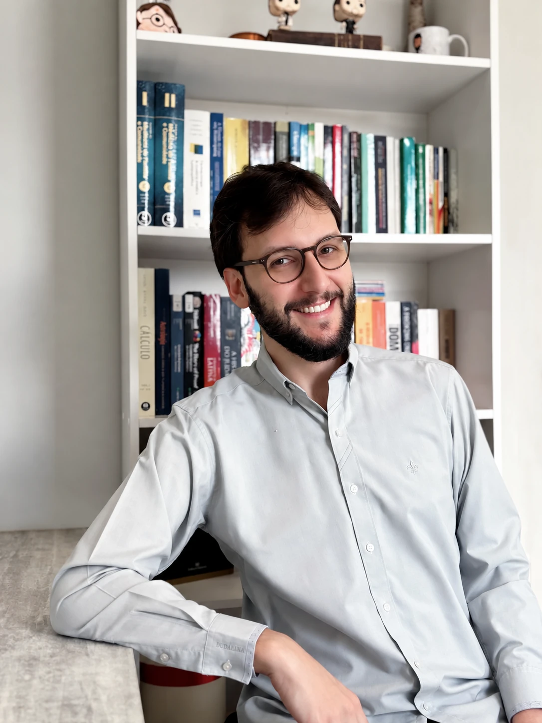 Iago Caires de óculos, sorrindo, sentado diante de sua estante de livros.